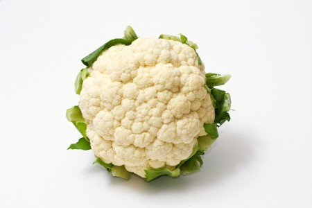 A full grown, round cauliflower with surrounding green leaves on a white background. The porous texture of the plant is clearly visible.