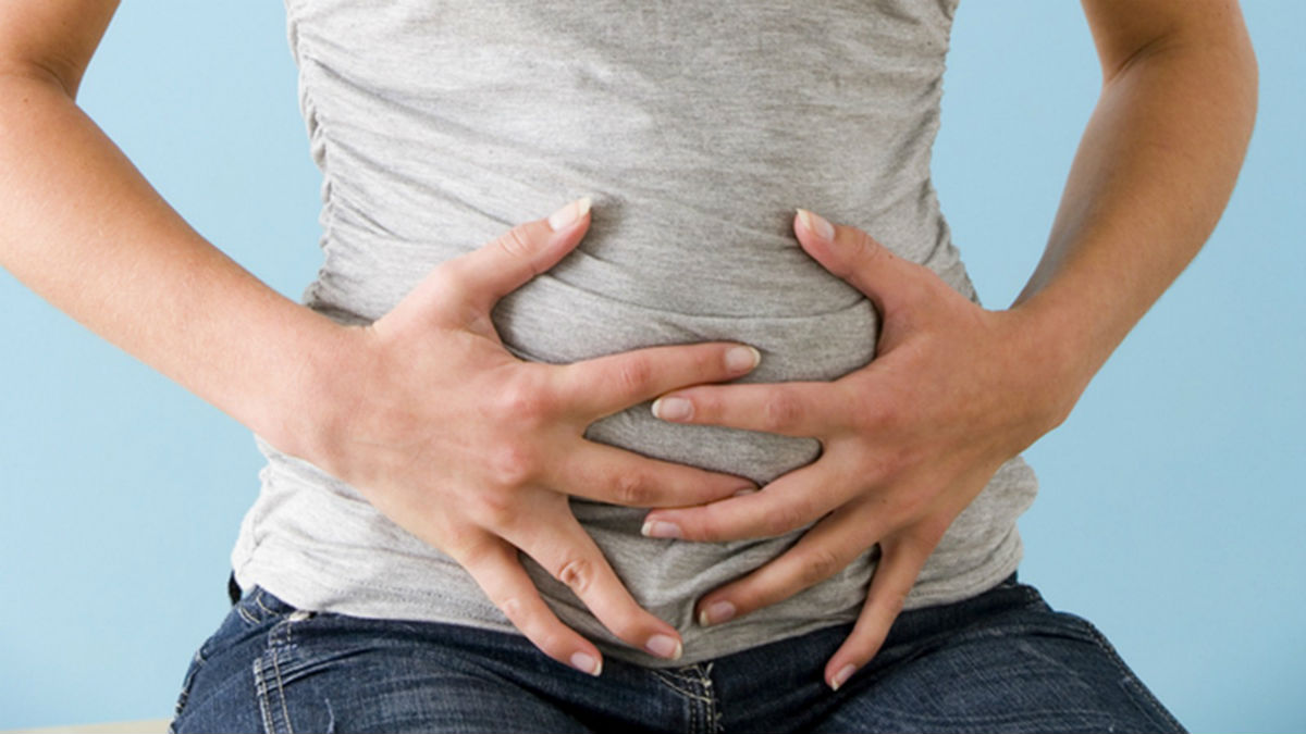  A woman in a gray shirt and blue jeans is holding her stomach in pain.